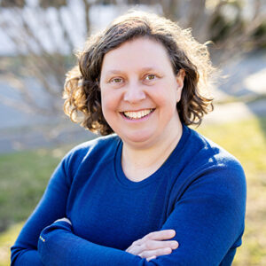 Photo of me, Leaf, in a blue shirt, smiling, with my arms crossed. I am a white woman with chin-length curly brown hair.