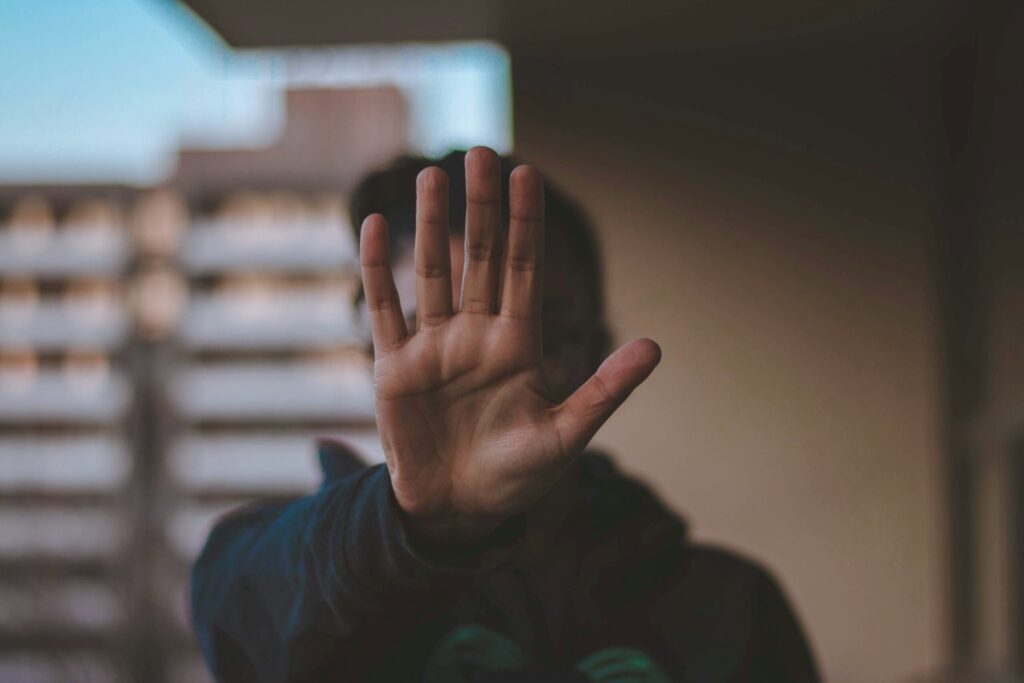 A person standing outside, holding up a hand in front of their face, with the palm of the hand facing outwards towards the viewer.