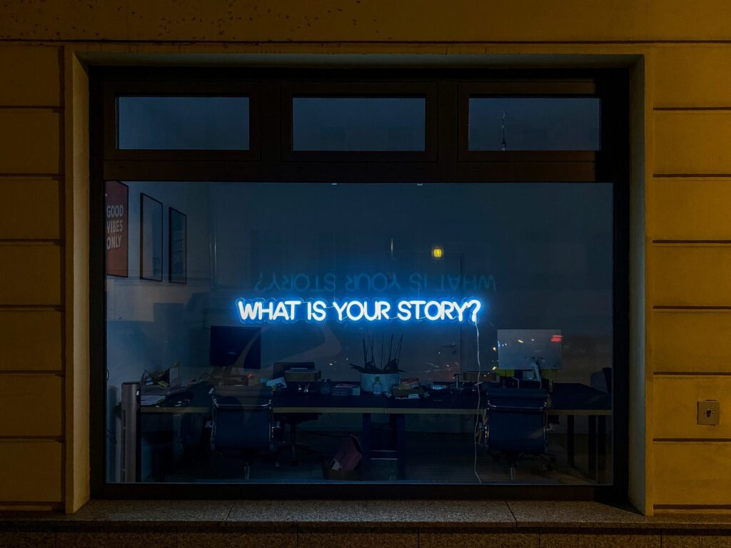 Neon sign that says "what is your story" glowing in an office window at night.