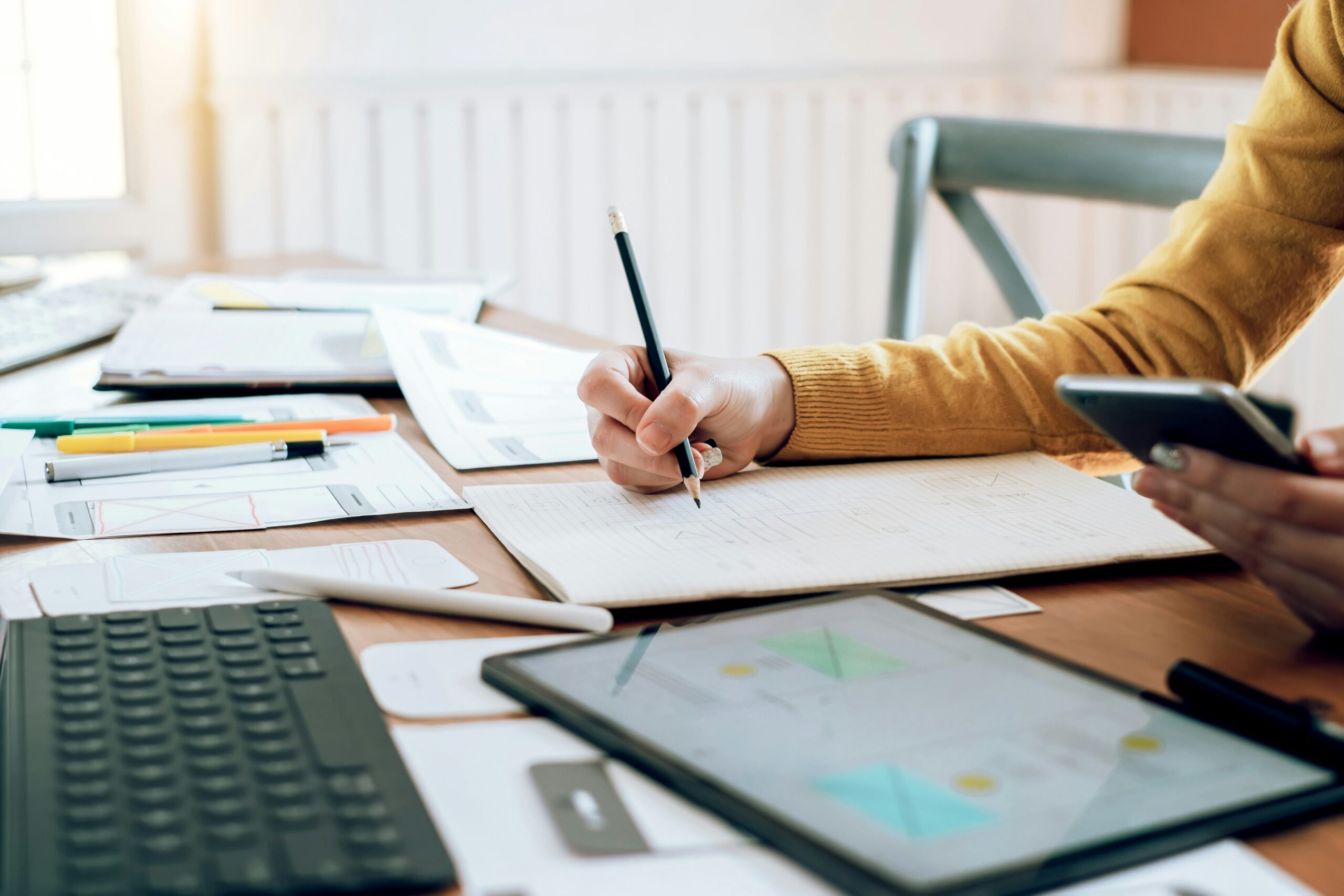A person holding a mobile device writes in a notebook on a table with other papers, pens, and a tablet, suggesting someone doing research.