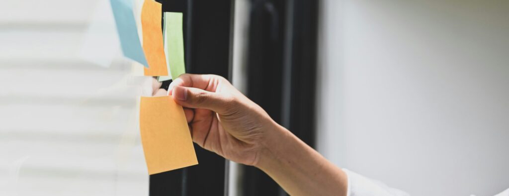 Someone holds a blank orange sticky note near three others that are stuck to a window